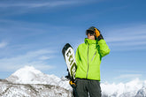 A man in a green jacket with a snowboard stands in front of a blue sky with a mountain range in the background