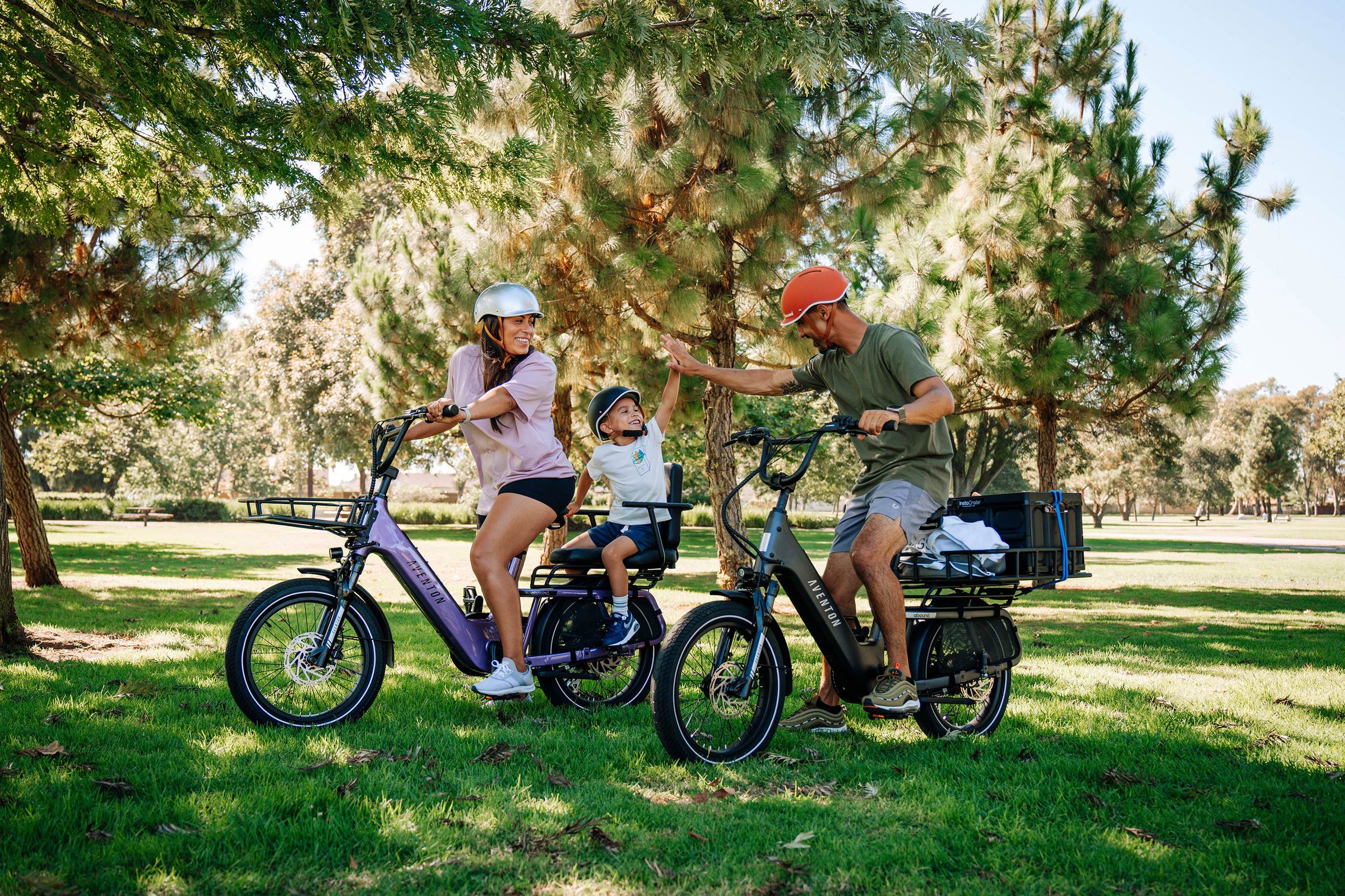 A family of riders on two cargo e-bikes in a sunny park, a child on the back seat high-fiving a parent.