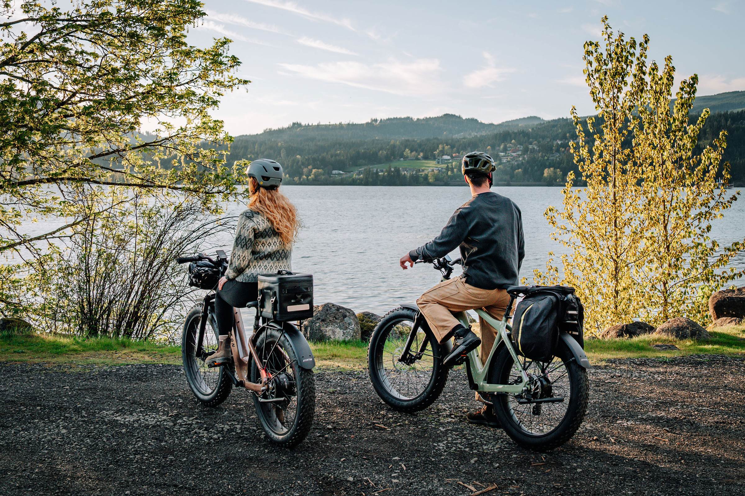 Two riders on e-bikes pausing on a lakeside path, looking out over the water and hills.